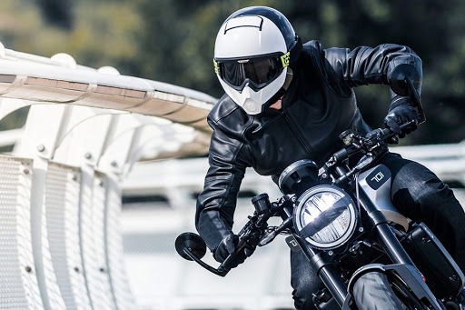 Pengendara motor dengan helm modular putih dan berjaket hitam yang sedang membelokkan motornya.